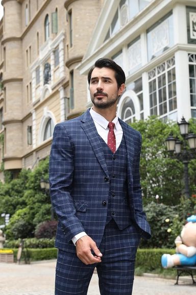Man in a checked suit walking outside near a building and greenery.