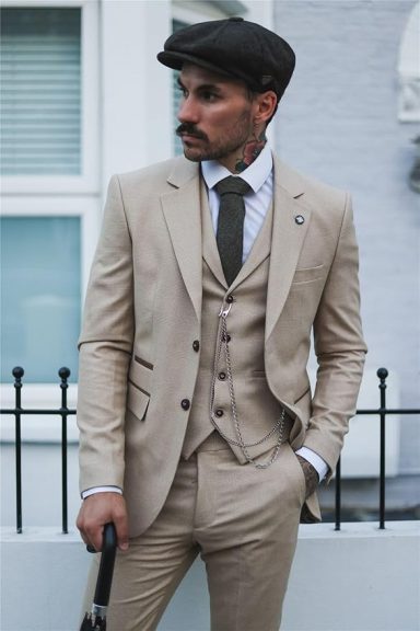 Man in a beige three-piece suit and flat cap, holding a cane, standing against a building.