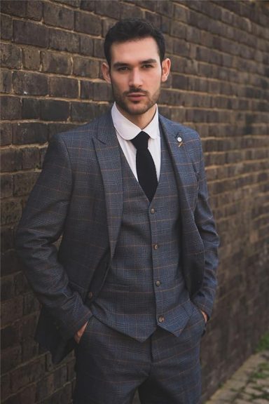 Man in a grey plaid suit and black tie standing against a brick wall.