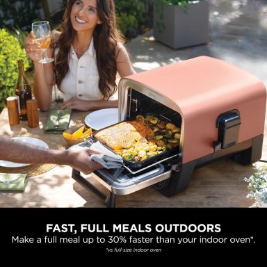 A woman smiles beside an outdoor cooking appliance with a tray of food.